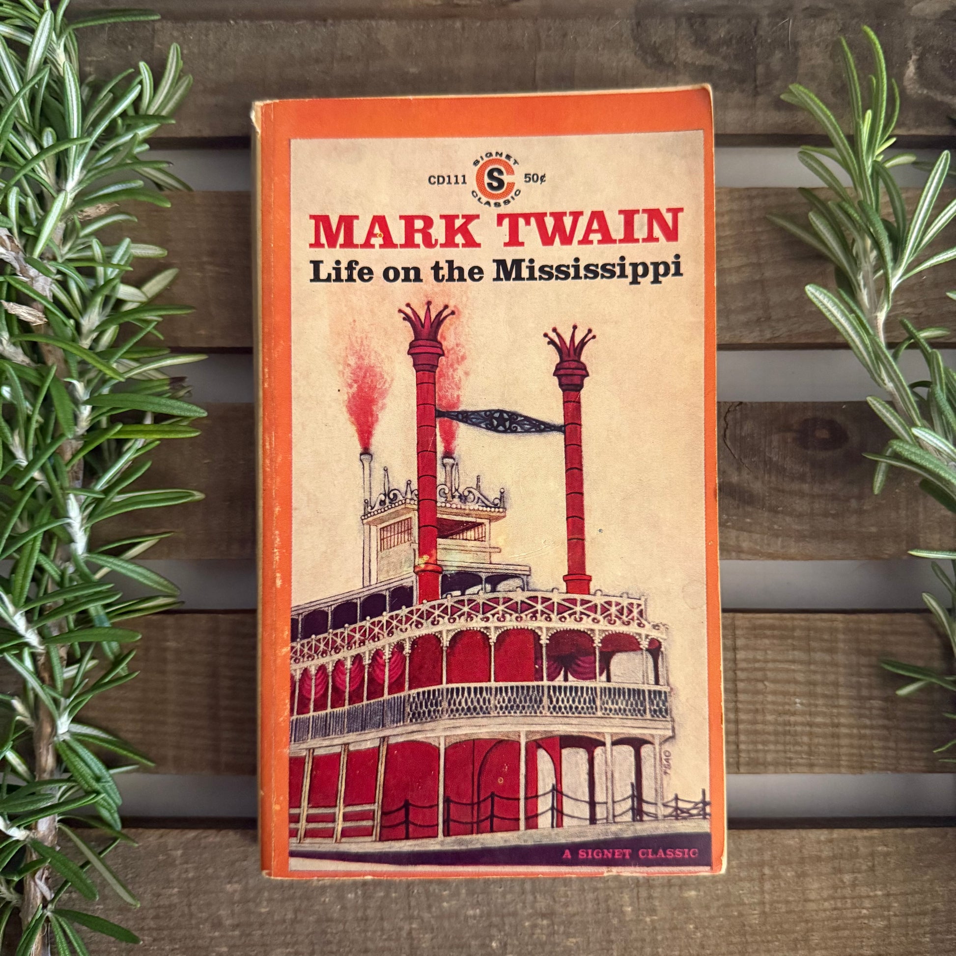 Book cover of 'Life on the Mississippi' by Mark Twain against a wooden background with plants.