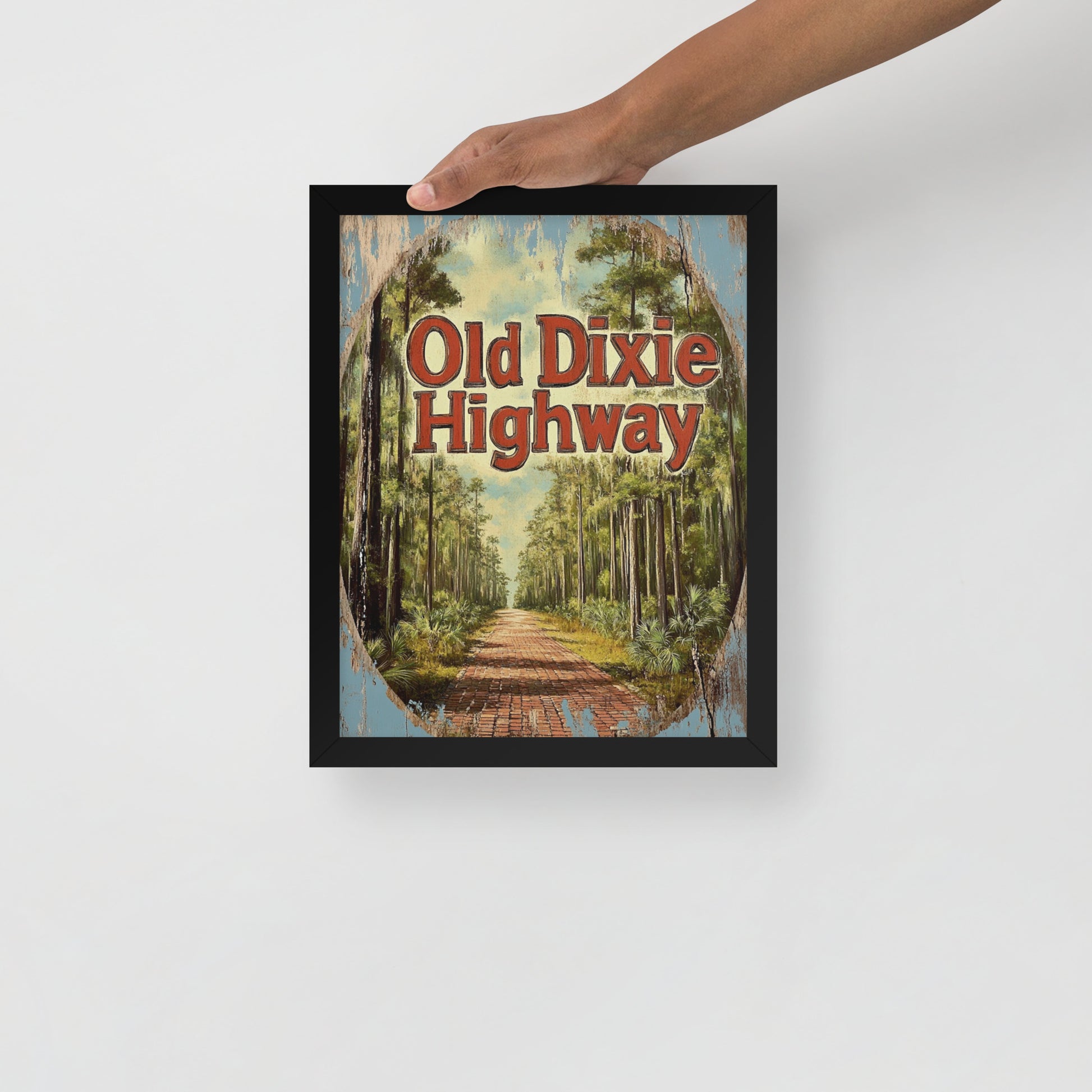 Framed artwork with 'Old Dixie Highway' text and forest scene, held by a hand on a white background.